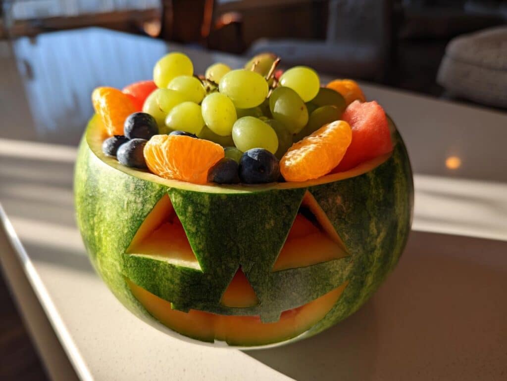 A carved watermelon in the shape of a Jack O Lantern, filled with a colorful assortment of grapes, blueberries, oranges, and watermelon pieces.