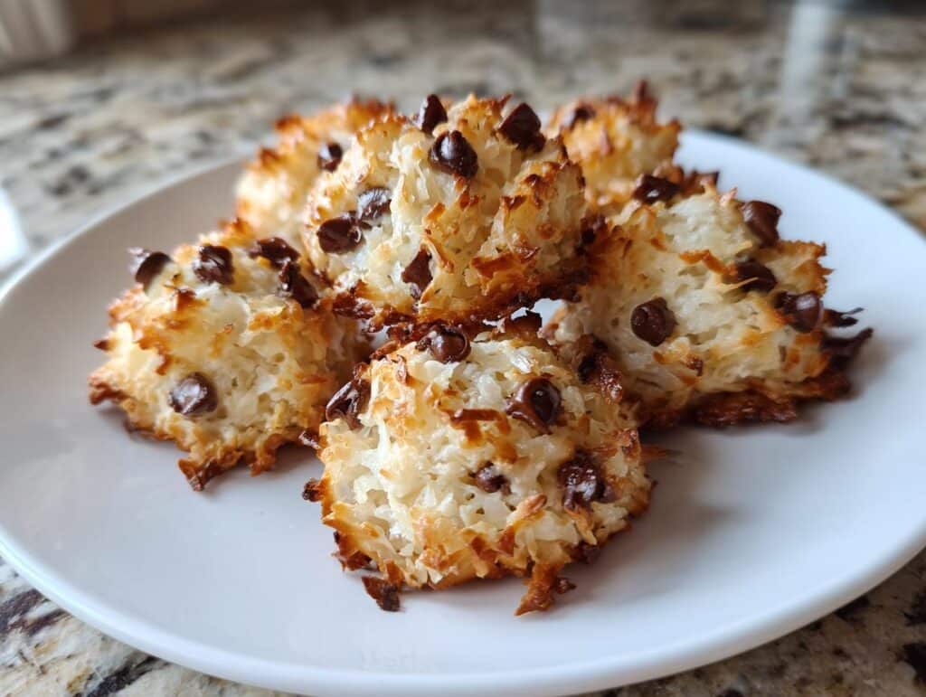 A close-up of several golden-brown Coconut Chocolate Chip Macaroons on a white plate, showing toasted coconut and melted chocolate chips.