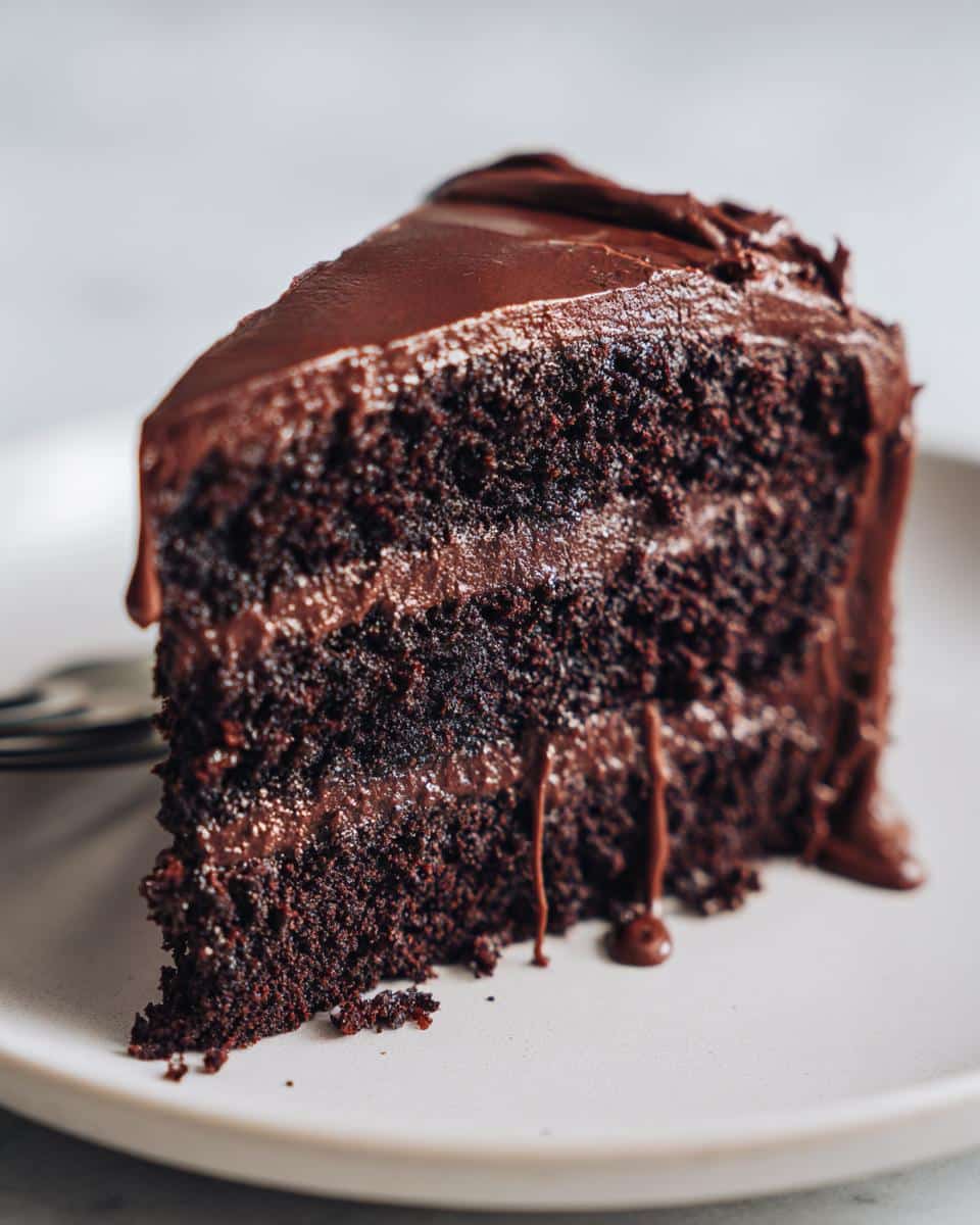 A close-up of a rich, dark slice of Death by Chocolate Cake with chocolate frosting and drips on a white plate.