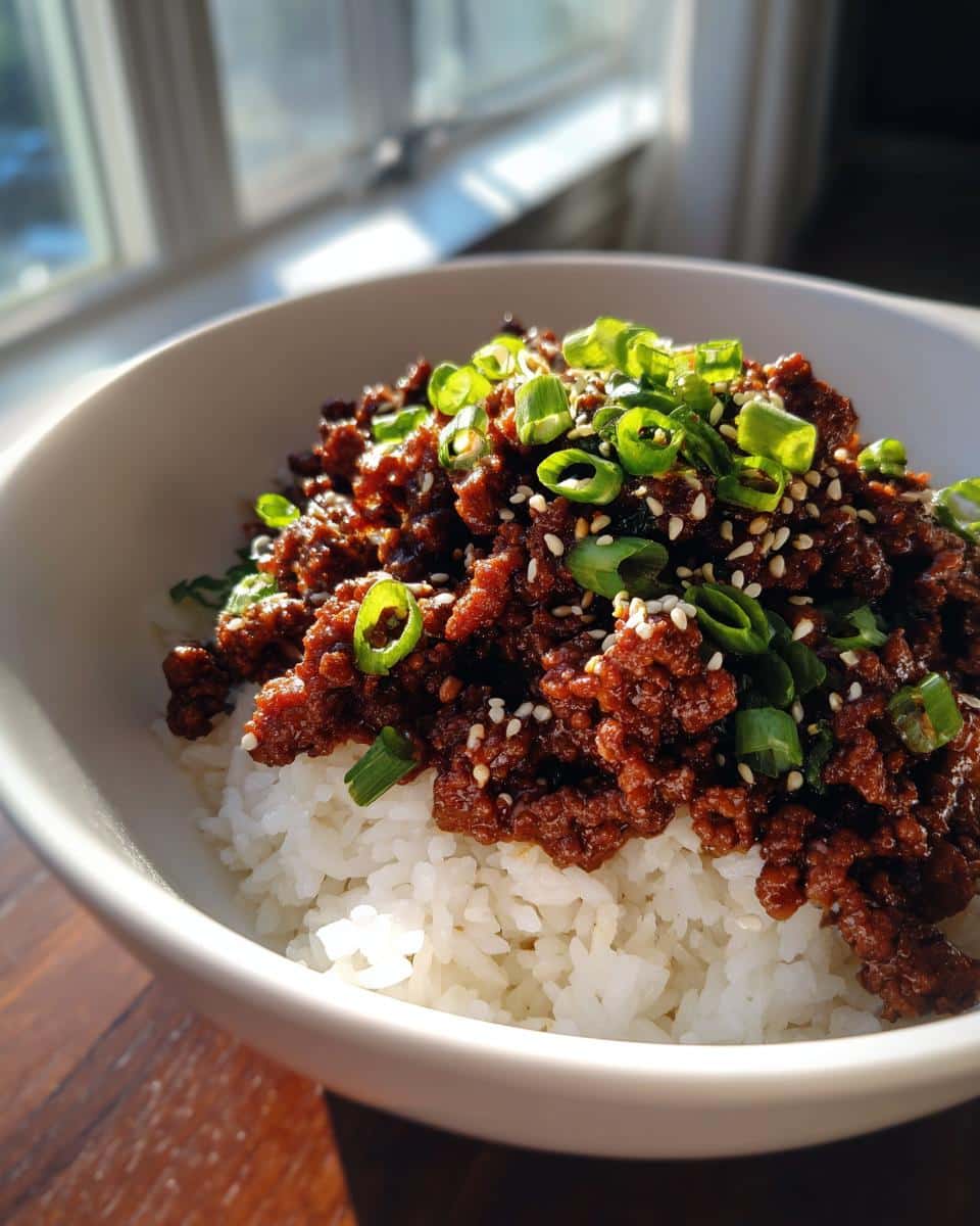 A close-up of a white bowl filled with white rice, topped with savory Korean Style Ground Beef Bowls, green onions, and sesame seeds.