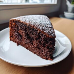 A close-up of a generous slice of The Most Amazing Chocolate Cake, dusted with powdered sugar, on a white plate.