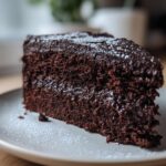Close-up of a moist slice of Rich Dreamy Home Made Chocolate Cake on a white plate, dusted with powdered sugar.